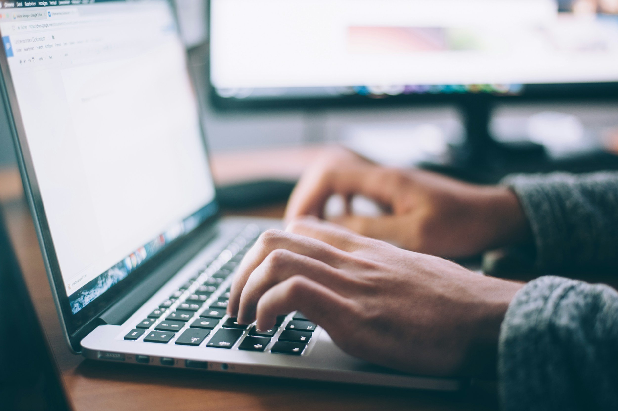 Person typing on a laptop with a monitor in the background.